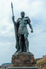 Large bronze statue of a muscular ancient warrior wearing helmet and cape, holding a long spear, standing on a rugged stone pedestal against a cloudy sky