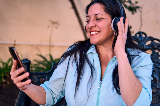 Beautiful, smiling, and cheerful Latina executive listening to music with headphones and her cell phone, relaxing and enjoying the day, sitting in a park. Learning, wellness, home office.