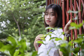 Japanese female student stands by red fence surrounded by greenery, looking thoughtful. Japanese schoolgirl