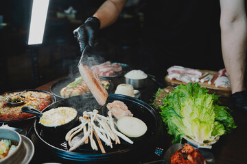 Sizzling Korean BBQ pork belly (samgyeopsal) on tabletop charcoal grill with rising smoke and traditional banchan side dishes, close-up shot in cozy restaurant setting with copy space.