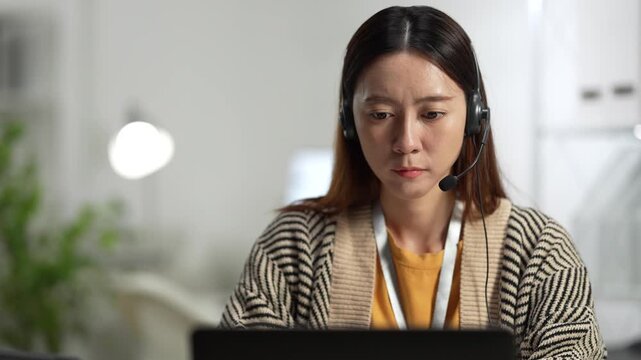 A professional Asian woman with a headset works on her laptop in a bright, modern office. She is focused on helping a customer or talking in an online meeting.