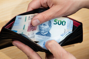 a male hand taking out a mexican five hundred pesos bill of a black leather wallet with a wooden...