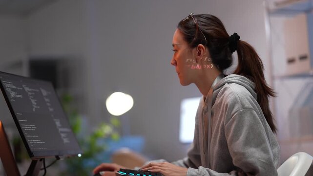 A smiling female programmer works at her computer workstation in a tech office. She is happy with her progress on a complex software development project after hours.