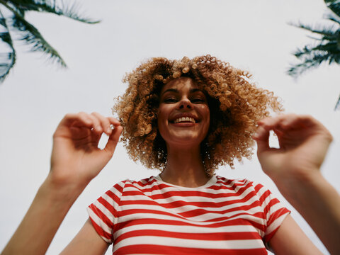 Low-angle shot of a joyful woman with curly hair, wearing a red and white striped shirt, smiling broadly against a bright sky and palm trees.