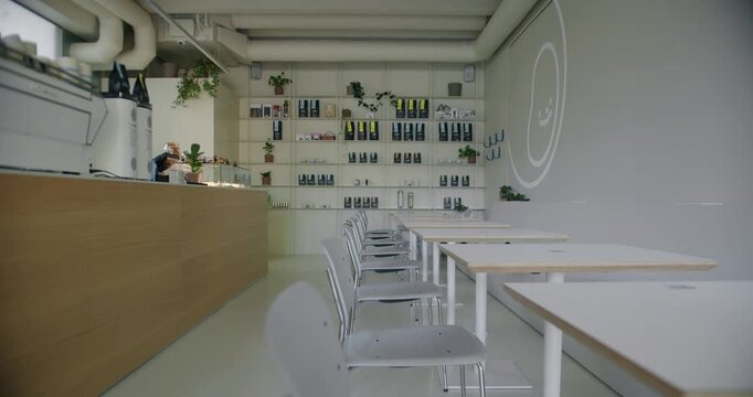 A spacious and well-lit cafeteria featuring a modern design with white chairs, wooden tables, and a barista station. The room is decorated with green plants and shelving filled with various items