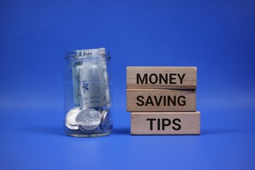 A jar containing coins and paper money sits beside wooden blocks stating 'Money Saving Tips,' implying financial responsibility and resourceful habits against an blue background.