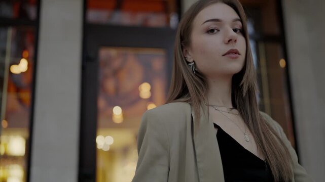 An image capturing an elegant young woman dressed in a stylish beige blazer, confidently posing in an urban, evening setting with soft lights in the background.