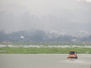 Fototapeta premium An orange machine boat is floating on the lake, designed to collect and clean water plants, with floating houses and bamboo structures in the background.