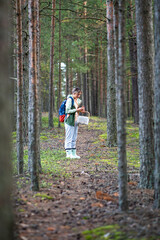 Female forager hold wild mushroom assessing it in pine woodland with basket among trees. Fungi identification in forest, edible verification, foraging hobby, pine thicket exploration on city break