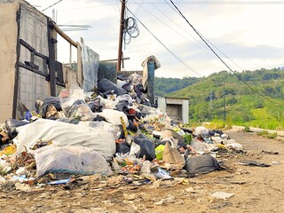 A large pile of garbage and debris sits on the side of a dirt road in a rural area, with hills in the background.