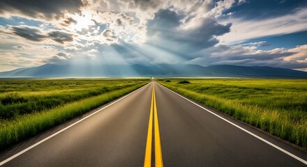 Fototapeta premium Straight Road Through Green Fields Towards Mountains Under a Dramatic Sky with Sunrays