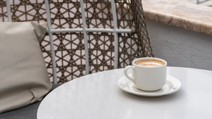 A white cup of coffee sits on a saucer on the table. Cappuccino foam is sprinkled with cinnamon. A cushion on a wicker swing chair. Resort cafe. Hotel. Cuba. Varadero.