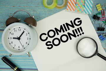 Coming Soon text on a paper surrounded by office supplies, a magnifying glass, and a clock on a...