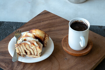A slice of cinnamon roll with white icing is on a small white plate with a fork, next to a white mug of black coffee on a wooden board.