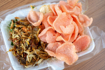 A top-down shot of a bowl of savory chicken porridge, topped with shredded chicken and large pink crackers, served in a takeaway container on a wooden surface.