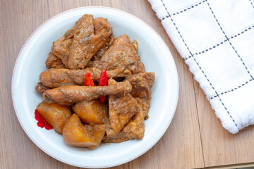 A plate of Indonesian braised chicken and potatoes in a brown sauce, resembling a chicken curry, garnished with red chili peppers, sits on a wooden table.