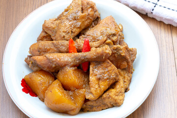 A plate of Indonesian braised chicken and potatoes in a brown sauce, resembling a chicken curry, garnished with red chili peppers, sits on a wooden table.