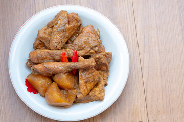 A plate of Indonesian braised chicken and potatoes in a brown sauce, resembling a chicken curry, garnished with red chili peppers, sits on a wooden table.