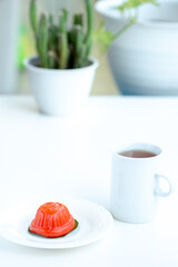 A small, red, tortoise-shaped Chinese sweet dumpling, known as ang ku kueh, sits on a white plate next to a white mug of tea. The background is a clean, bright, out-of-focus scene with a potted cactus
