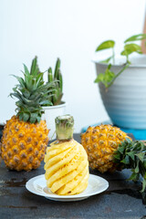 A bright, high-key photo of a peeled pineapple (Ananas comosus) with a spiral cut. The fresh, juicy fruit sits on a white plate, with whole pineapples and green plants creating a soft bokeh background