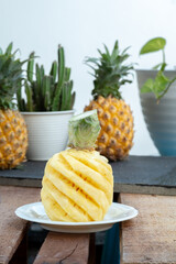 A fresh, spirally-cut pineapple (Ananas comosus) sits on a plate atop a rustic wooden crate. In the bright, soft-focus background are whole pineapples and potted plants, creating a natural still life.