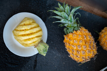 A whole ripe pineapple with its spiky skin is next to a peeled, carved pineapple on a white plate,...