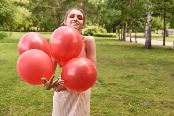 Smiling young caucasian female holding red balloons in a park on a sunny day.