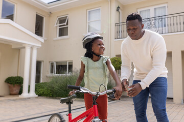 Father teaching daughter to ride bicycle in driveway, both smiling and engaged