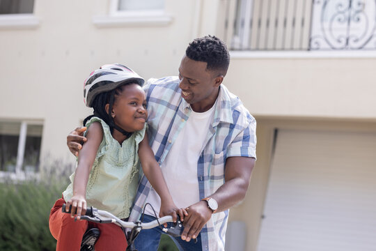 African American father teaching daughter to ride bicycle, both smiling outdoors