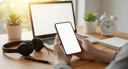 Person holding a smartphone with a laptop and headphones on a desk