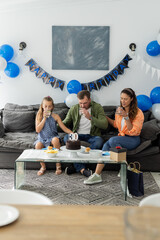 Family celebrating birthday at home, enjoying cake and drinks on sofa