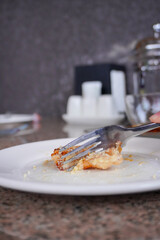 Hand holding fork with small food portion on white plate, with blurred table setting in background.