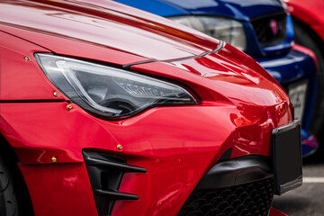 A detailed, close-up shot of the front of a custom red sports car, featuring the sleek headlight and wide body kit with riveted fenders. A blue car is partially visible in the blurred background.