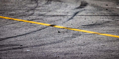 Close-up of a textured asphalt road surface marked with a faded yellow line and numerous black tire...
