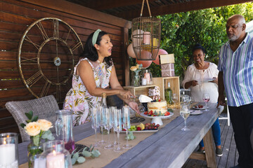 Family celebrating birthday outdoors, woman cutting cake while others enjoy wine