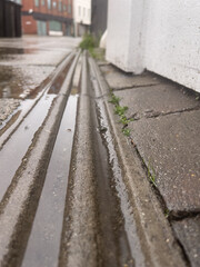 The image captures a dreary urban alley, its wet pavement glistening with recent rain, reflecting muted tones of grey and brown. Parallel grooves in the concrete channel shallow puddles, bordered by a