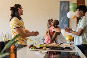 Friends celebrating at home, hugging and clapping around kitchen table with snacks