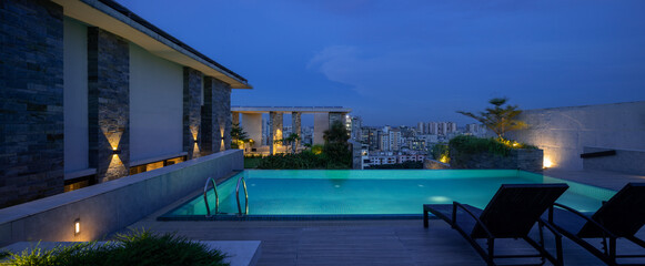 A stunning panoramic view of a luxurious rooftop pool at dusk. The tranquil, turquoise water is illuminated from within, complementing the warm light from the wall sconces. 