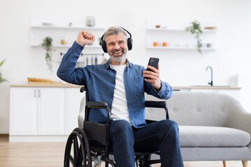 Mature Caucasian man in wheelchair using phone, listening to music with headphones, smiling and celebrating. Modern home setting with cozy interior. Concept of joy music and accessibility enhancement.