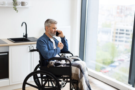 Senior Caucasian man sitting in wheelchair by kitchen indoors using phone near window. Scene depicts everyday lifestyle, accessibility, comfort, connection, communication, independence, and inclusion