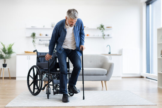 Disabled Caucasian male, age around 50s, practicing walking with cane support near wheelchair indoors. Bright living room setting conveys recovery and determination.