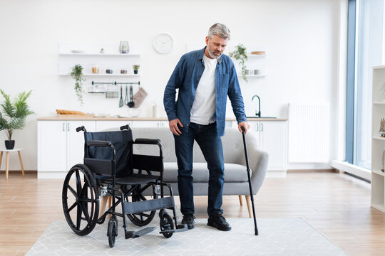 Disabled adult man stands with cane near wheelchair indoors. Concept of recovery and mobility support. - Powered by Adobe