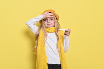 Photo of a funny impressed little girl with a hat and a hat. holding a thermometer in autumn, it feels bad on a yellow background
