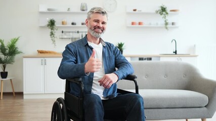 Senior Caucasian man seated in wheelchair showing thumbs-up gesture expressing positivity indoors. Image showcases accessibility, mobility aid, and cheerful demeanor in modern home environment.