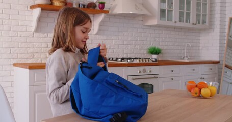 Happy elementary school kid getting ready for school in kitchen, packing backpack with books and supplies. Excited smiling child feeling confident and motivated before starting new day of learning.