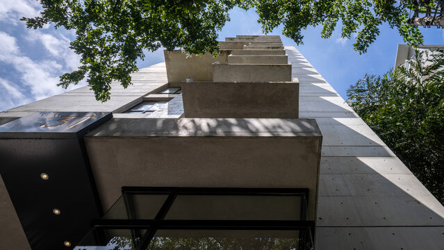 Low-angle shot of a modern facade with raw concrete balconies framed by green trees. Bright sunlight highlights the brutalist-inspired design.