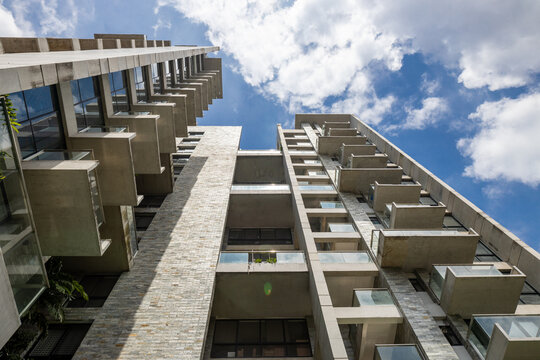 Striking upward shot of a high-rise building with concrete and stacked stone walls and alternating balconies. A blue sky with clouds creates a dramatic backdrop.
