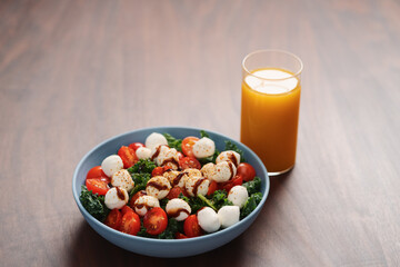 Blue bowl of salad with mozzarella, cherry tomatoes and kale, seasoned with balsamic sauce on walnut wood table with glass of orange juice
