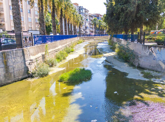 Fototapeta premium Urban river with dirty water, algae, and scattered litter in a modern city. Environmental pollution problem caused by urbanization and lack of waste management. Concept of climate change, water