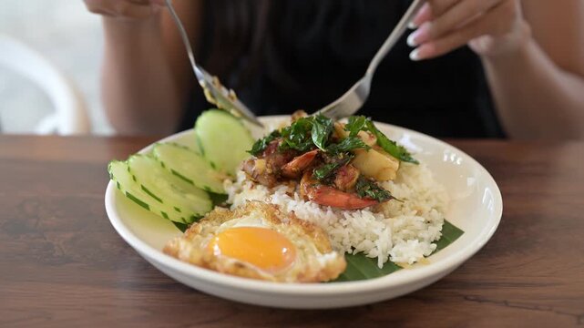 Close-up of woman in restaurant eating Thai holy basil seafood dish with fried egg and rice during day